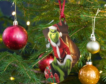 Puede incluir: Adorno navideño con forma de calcetín, con el Grinch vestido de Papá Noel. El adorno es verde, rojo y blanco, con una cinta roja para colgar. El fondo muestra una chimenea y regalos.