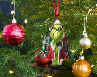 Puede incluir: Un adorno navideño con el Grinch con un abrigo rojo de Papá Noel con ribete de piel blanca. El adorno cuelga de una cinta roja y está rodeado de otros adornos navideños y ramas de árbol. El Grinch sonríe con picardía.