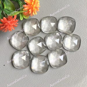 May include: Ten clear, faceted, square-shaped gemstones. The gemstones are arranged in a random pattern on a gray surface.