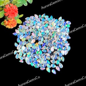 May include: A collection of numerous pear-shaped opals displaying iridescent flashes of blue, green, and white light. The gemstones are arranged in a cluster, with the text AuroraGemsCo visible in the background. Two orange flowers are at the top.