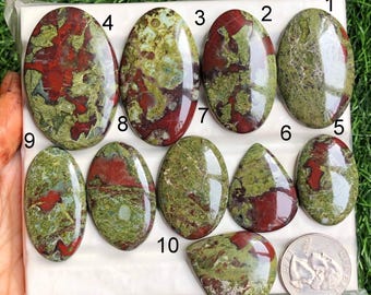 May include: A collection of ten polished gemstones in various oval and teardrop shapes. The stones feature a mix of red and green hues, with a coin for size comparison. The gemstones are displayed on a white surface.