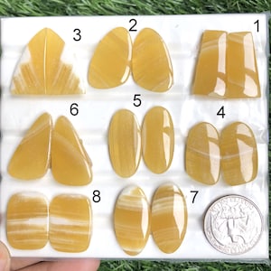 May include: A collection of eight pairs of polished, yellow gemstones in various shapes, including ovals, teardrops, and rectangles. Some stones have white veining. A quarter coin is included for size reference.