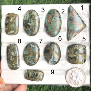 Puede incluir: Colección de nueve cabujones pulidos de varias formas, incluyendo ovalados, rectangulares y triangulares. Las piedras presentan un patrón de matriz verde y dorado, con una moneda para comparar el tamaño. Los cabujones se muestran sobre una superficie blanca.