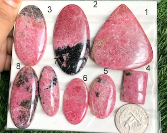 May include: Assortment of polished rhodochrosite gemstones in various shapes, including oval, triangular, and rectangular. The stones display a range of pink hues with black inclusions. A US quarter coin is included for size reference.