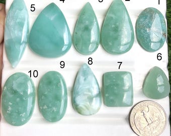 May include: A collection of ten polished gemstones in various shapes and shades of green and blue, including oval, teardrop, and rectangular forms. A US quarter is included for size reference. The gemstones are displayed on a white surface.