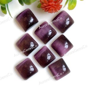 May include: Eight purple square-shaped cabochon gemstones with a chatoyant effect. The gemstones are arranged in a circular pattern on a white surface.
