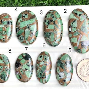 Puede incluir: Ocho piedras preciosas ovaladas con un patrón de mosaico de secciones verdes, negras y color cobre. Se incluye una moneda para comparar el tamaño. Las piedras preciosas se muestran sobre una superficie blanca.