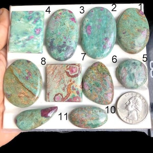 Op de afbeelding: Een verzameling van elf gepolijste edelstenen in verschillende vormen, waaronder ovalen, rechthoeken en vrije vormen. De stenen hebben een mix van teal, groen en roze tinten, met een munt voor maatvergelijking. De edelstenen worden gepresenteerd op een wit oppervlak.