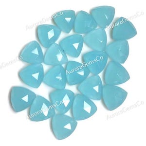 May include: A collection of light blue, triangular gemstones with faceted surfaces. Each gem has a triangular facet in the center. The gems are scattered across a white surface. The text "AuroraGemsCo" is printed on the image.