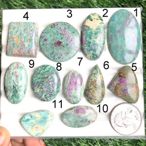 May include: A collection of eleven polished ruby in zoisite gemstones in various shapes, including oval, round, and teardrop. The stones display a mix of green, pink, and red hues. A US quarter is included for size reference.