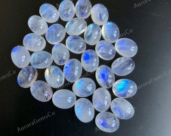 May include: A collection of oval-shaped moonstone cabochons, with a milky white color and iridescent blue and green flashes. The stones are arranged in a loose pile on a black background.