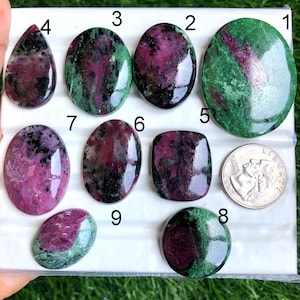 Puede incluir: Una colección de piedras preciosas de rubí zoisita pulidas en varias formas, incluyendo ovaladas, en forma de lágrima y redondas. Las piedras muestran una mezcla de tonos rojo intenso y verde, con una moneda para comparar el tamaño. Las piedras preciosas se exhiben sobre una superficie blanca.