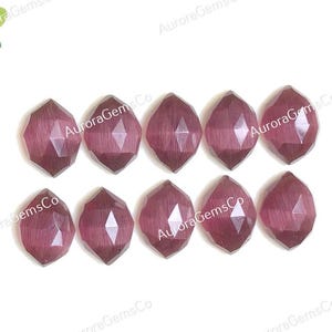 May include: Ten oval-shaped, faceted gemstones in a deep reddish-purple colour are displayed on a white surface. The gems have a symmetrical, geometric cut, reflecting light. The image includes the text "Aurora Gems Co" in the background.