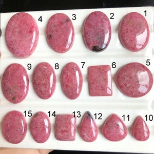 May include: A collection of fifteen polished pink rhodonite cabochons arranged in three rows on a white display tray. Each stone is numbered from 1 to 15 and features varying shapes including ovals, rectangles, and teardrops with unique patterns.