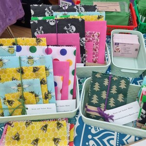 Puede incluir: Variedad de envoltorios de alimentos reutilizables de cera de abeja con diversos diseños, incluyendo abejas, lunares, calaveras y motivos florales. Los envoltorios están ordenados en pequeños recipientes verde claro. Algunos están atados con hilo o cuerda morada.