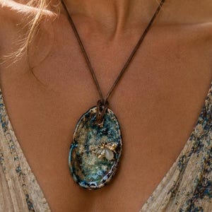 May include: A necklace featuring an oval-shaped pendant with a mottled blue and green finish, adorned with a small metallic bee emblem. The pendant hangs from a thin brown cord, resting against a cream-colored garment with a blue floral pattern.