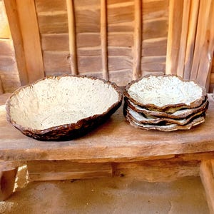 Pu&ograve; includere: Una grande ciotola in ceramica strutturata e una pila di piatti pi&ugrave; piccoli con un design simile. Le ciotole e i piatti hanno un bordo ruvido e organico e un interno screziato bianco sporco, con esterno marrone scuro.