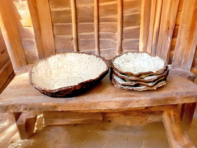 Pu&ograve; includere: Una grande ciotola in ceramica strutturata e una pila di piatti pi&ugrave; piccoli con un design simile. Le ciotole e i piatti hanno un bordo ruvido e organico e un interno screziato bianco sporco, con esterno marrone scuro.