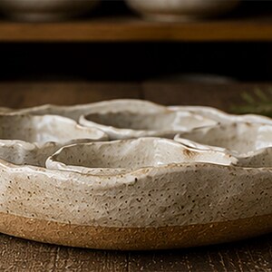 May include: A round, beige ceramic serving platter with a textured, speckled surface and an uneven, wavy rim. The platter contains three smaller, matching ceramic bowls with similar irregular edges, all featuring a light, speckled glaze finish.