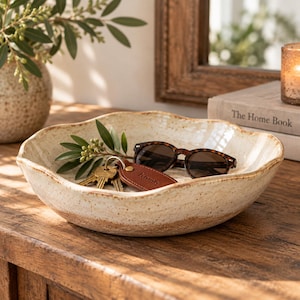 May include: A shallow, round ceramic bowl with a speckled cream glaze and a wavy, uneven rim. The bowl features a raw, unglazed terracotta-colored base and contains a pair of tortoiseshell sunglasses and a brown leather keychain with the word home.