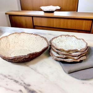 Pu&ograve; includere: Una grande ciotola strutturata bianco sporco con bordo marrone &egrave; accanto a una pila di piatti pi&ugrave; piccoli abbinati. Un tovagliolo grigio &egrave; piegato vicino. La ceramica fatta a mano ha un'estetica rustica e organica.