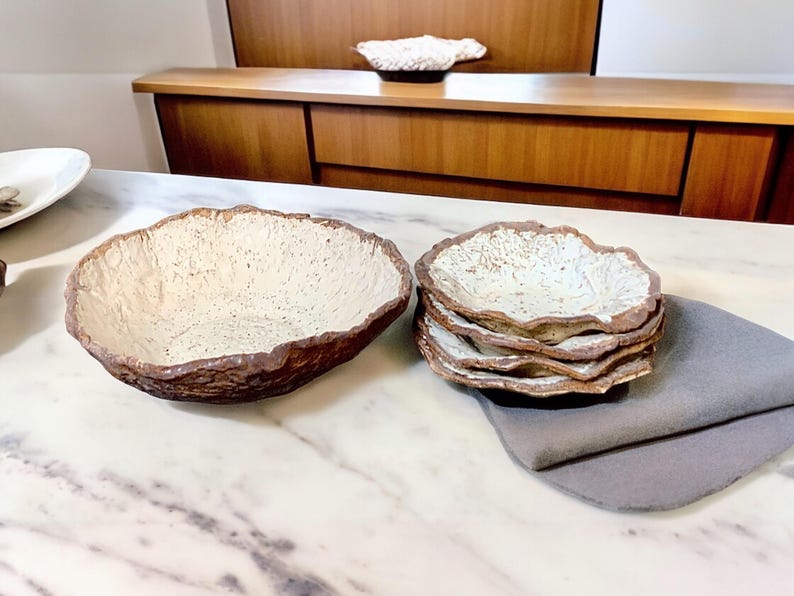 Pu&ograve; includere: Una grande ciotola strutturata bianco sporco con bordo marrone &egrave; accanto a una pila di piatti pi&ugrave; piccoli abbinati. Un tovagliolo grigio &egrave; piegato vicino. La ceramica fatta a mano ha un'estetica rustica e organica.