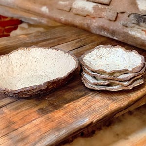 Pu&ograve; includere: Una ciotola rustica in ceramica e diversi piattini con interno strutturato bianco sporco e bordo marrone dalla forma organica. Gli oggetti sono disposti su una superficie di legno invecchiato, con una candela accesa sullo sfondo.