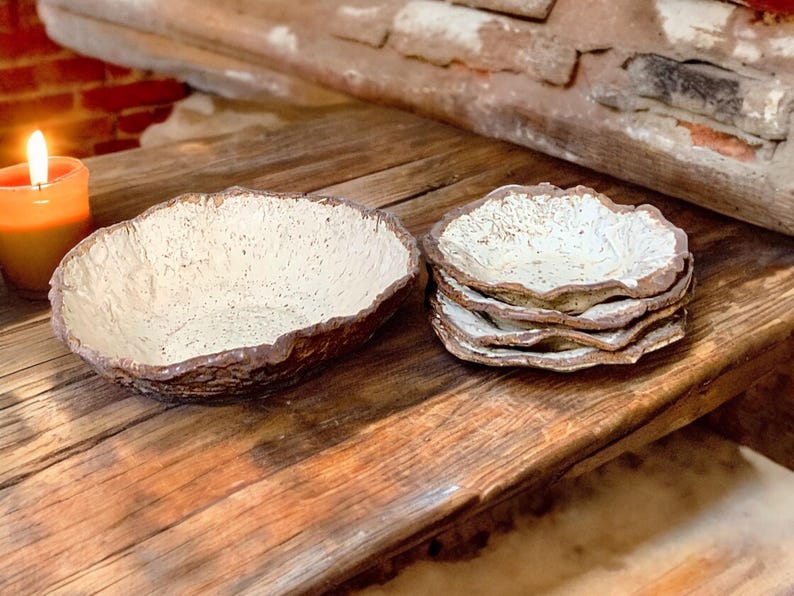 Pu&ograve; includere: Una ciotola rustica in ceramica e diversi piattini con interno strutturato bianco sporco e bordo marrone dalla forma organica. Gli oggetti sono disposti su una superficie di legno invecchiato, con una candela accesa sullo sfondo.
