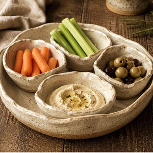 May include: A set of four speckled, off-white ceramic bowls with irregular, organic edges arranged on a matching circular tray. The bowls contain fresh orange baby carrots, green celery sticks, green olives, and a creamy hummus dip with oil.