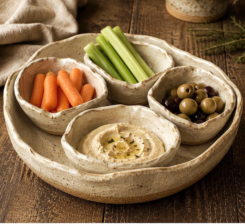 May include: A set of four speckled, off-white ceramic bowls with irregular, organic edges arranged on a matching circular tray. The bowls contain fresh orange baby carrots, green celery sticks, green olives, and a creamy hummus dip with oil.