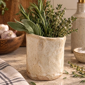 May include: A cylindrical ceramic utensil holder with a cream-colored, speckled, and textured finish. The vessel is filled with a bundle of fresh green herbs, including sprigs of rosemary, sage leaves, and flowering thyme stems.