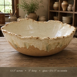 May include: A large, round ceramic bowl with a textured, off-white interior and a raw, tan-colored exterior base. The rim is uneven and brown-speckled. The bowl measures 13.5 inches across, 5 inches deep, with a 2.5 to 3-inch glaze on the outside.