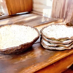 Pu&ograve; includere: Una grande ciotola dalla forma irregolare e una pila di piatti abbinati. I pezzi di ceramica hanno un esterno marrone strutturato e un interno bianco sporco macchiettato. Le stoviglie rustiche sono esposte su una superficie di legno.