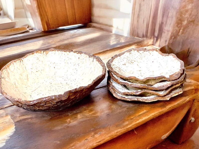 Pu&ograve; includere: Una grande ciotola dalla forma irregolare e una pila di piatti abbinati. I pezzi di ceramica hanno un esterno marrone strutturato e un interno bianco sporco macchiettato. Le stoviglie rustiche sono esposte su una superficie di legno.