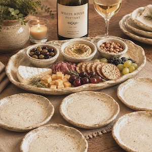 May include: A set of speckled beige ceramic tableware including a large oval serving platter with wavy edges, five smaller round plates, and three small bowls. The platter holds an assortment of cheese, crackers, grapes, olives, and hummus.