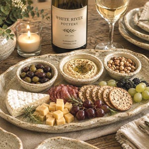 May include: A large, oval, speckled beige ceramic serving platter with wavy edges, holding three small matching bowls filled with olives, hummus, and nuts, alongside a spread of brie cheese, cubed cheddar, salami, crackers, and grapes.