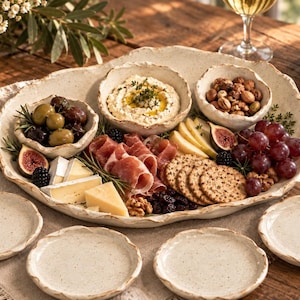 May include: A large, irregular, off-white ceramic serving platter with a speckled finish and wavy edges, containing two smaller matching bowls filled with dip and nuts, surrounded by prosciutto, cheese, crackers, grapes, figs, and olives.