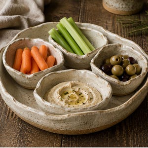 May include: A set of four off-white speckled ceramic bowls arranged on a matching circular tray. The bowls contain orange baby carrots, green celery sticks, green and black olives, and a smooth beige dip topped with olive oil and herbs.