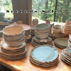 May include: A table display of Seconds Sale pottery, featuring stacks of light blue and white ceramic bowls and plates with a rough, textured finish.