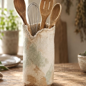 May include: A cylindrical ceramic utensil holder with a textured, uneven rim, featuring a mottled finish in shades of off-white, sage green, and tan. The holder contains three wooden kitchen spoons and a silver-toned metal wire whisk.