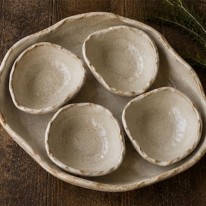 May include: A set of five ceramic pieces consisting of one large, irregularly shaped serving platter and four smaller, matching bowls. Each piece features an off-white, speckled glaze with a textured, uneven rim finished in a light brown tone.