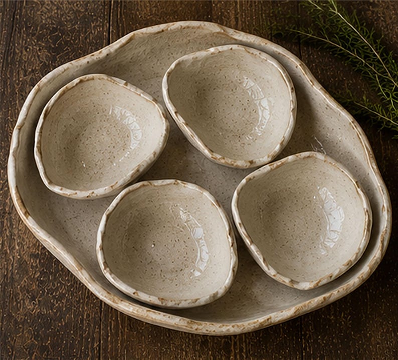 May include: A set of five ceramic pieces consisting of one large, irregularly shaped serving platter and four smaller, matching bowls. Each piece features an off-white, speckled glaze with a textured, uneven rim finished in a light brown tone.