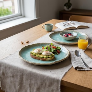 May include: A breakfast table setting with a light blue ceramic plate holding avocado toast topped with poached eggs and a side salad. A matching bowl with berries, a mug, and a glass of orange juice are also present. A linen runner and wooden table complete the scene.