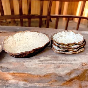 Pu&ograve; includere: Una collezione di ciotole e piatti in ceramica fatti a mano. Le ciotole hanno un interno strutturato bianco sporco e un bordo marrone scuro. &Egrave; visibile anche una pila di piatti con un design simile, il tutto su una superficie di legno rustico.