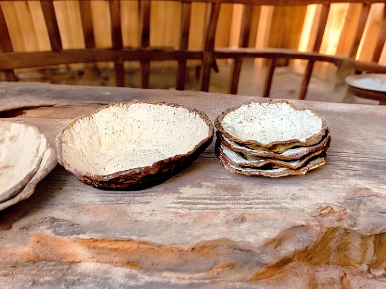 Pu&ograve; includere: Una collezione di ciotole e piatti in ceramica fatti a mano. Le ciotole hanno un interno strutturato bianco sporco e un bordo marrone scuro. &Egrave; visibile anche una pila di piatti con un design simile, il tutto su una superficie di legno rustico.