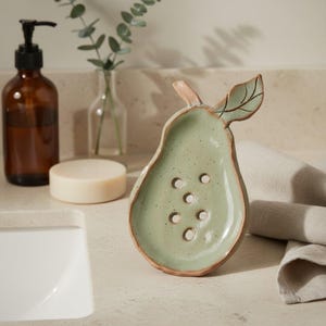May include: A pear-shaped ceramic soap dish in a light sage green glaze with a speckled finish and a brown rim. The dish features a sculpted stem and leaf detail at the top, along with five circular drainage holes in the center of the basin.
