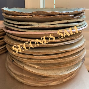 May include: A tall stack of round ceramic plates with irregular, wavy edges. The plates feature a speckled texture in shades of light blue, beige, and soft pink. The text SECONDS SALE is overlaid in gold lettering across the center of the stack.