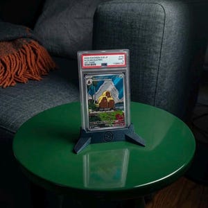 Pode incluir: Uma carta colecionável Pokemon SV8 JP Alolan Dugtrio Art Rare de 2024, graduada pela PSA em um estojo de plástico transparente, exibida em um suporte de plástico azul escuro. A carta apresenta uma ilustração colorida de três criaturas marrons.