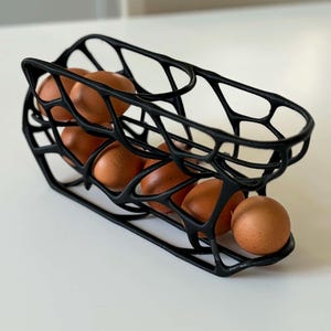 Könnte beinhalten: Ein schwarzer, 3D-gedruckter Eierhalter mit einer organischen, netzartigen Gitterstruktur. Der längliche, offene Rahmenbehälter fasst sechs braune Hühnereier, die in einer Reihe angeordnet sind und ein modernes, geometrisches Design aufweisen.