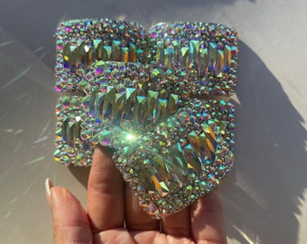 May include: A set of four iridescent, square-shaped, rhinestone-covered buckles. The buckles are made of metal and have a shiny, reflective finish.